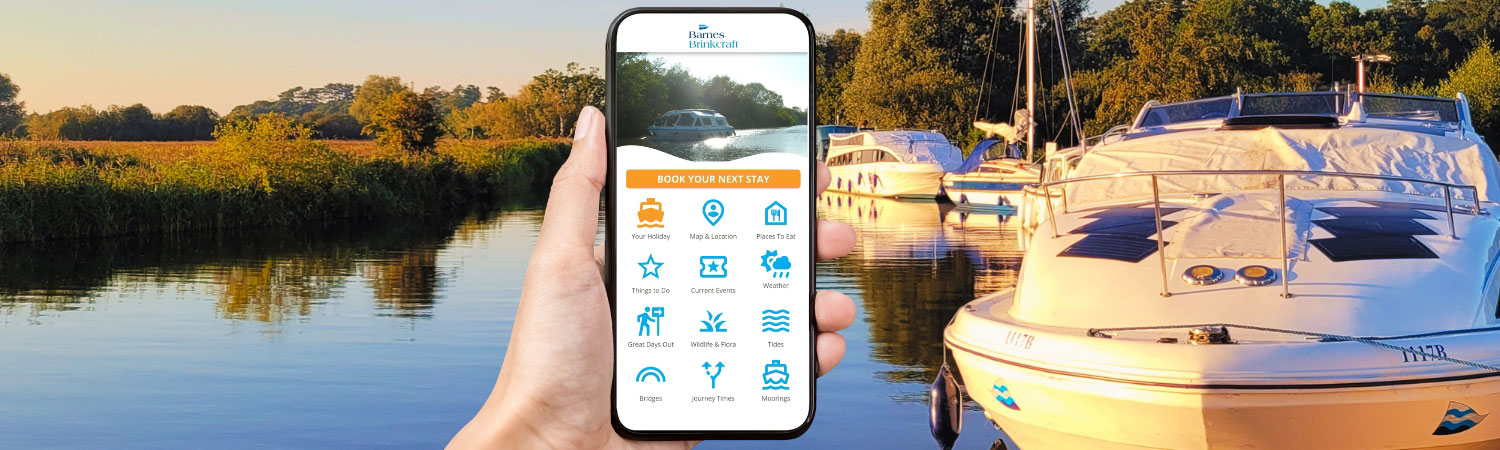 A hand holding a mobile phone, showing the homepage of the Barnes Brinkcraft App. Broads cruiser boats moored in background
